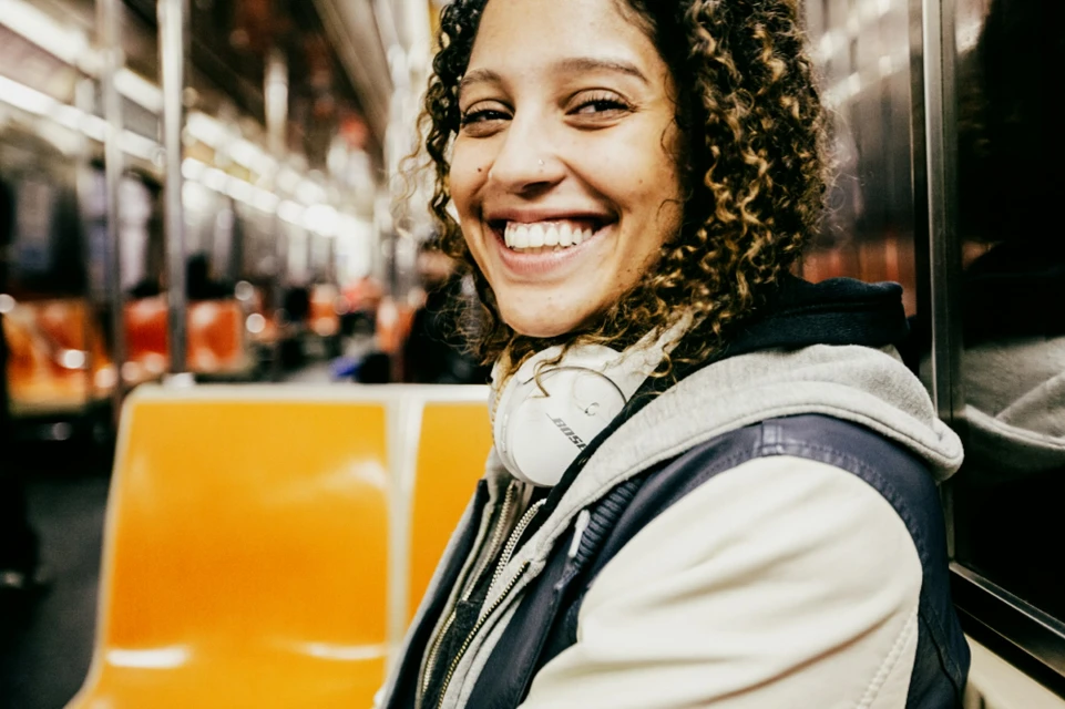 A smiling young woman with curly hair, wearing headphones, is photographed on a subway train with orange seats and steel accents.