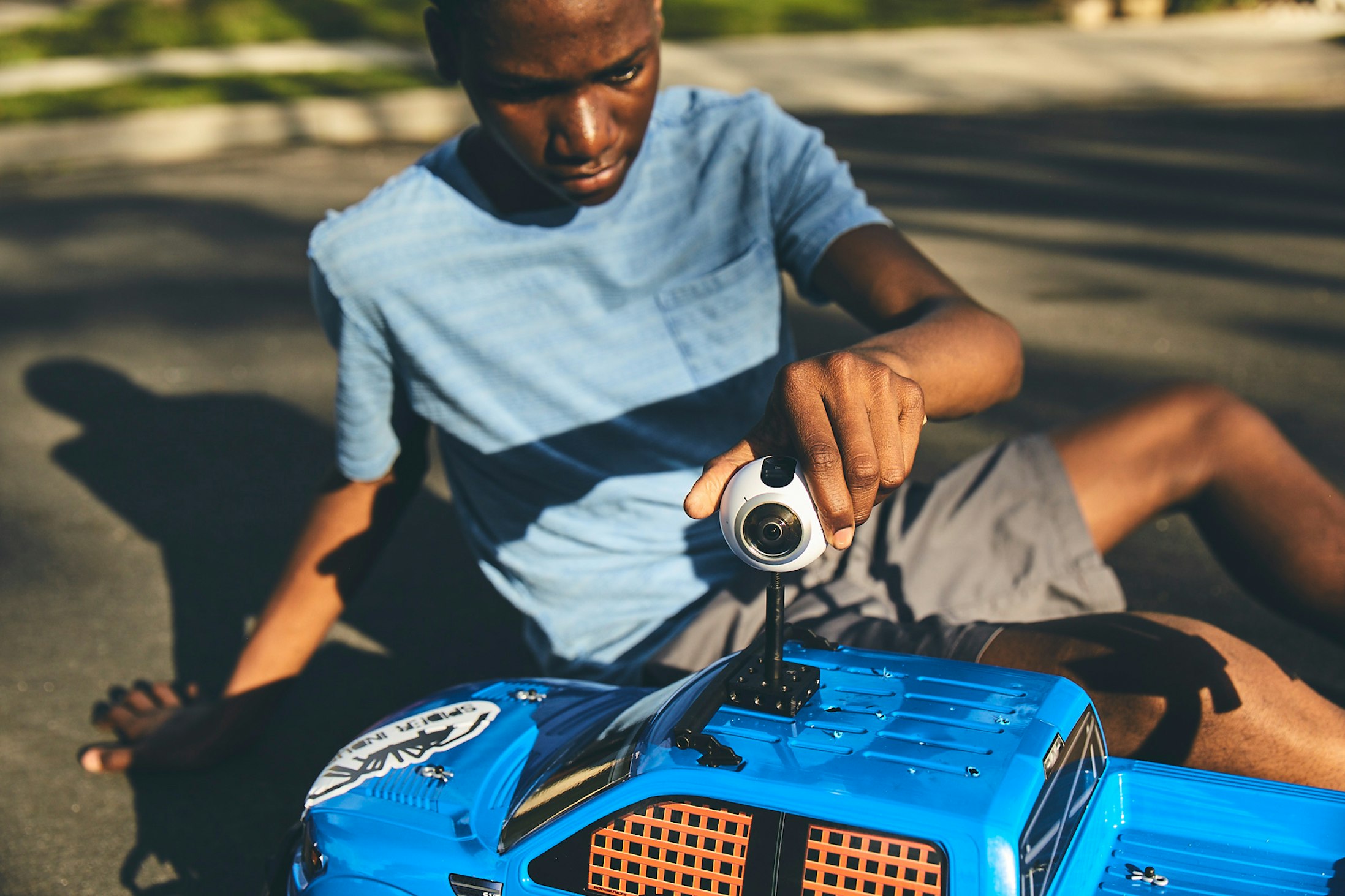 Teenage boy sits outdoors on pavement next to a blue toy truck holding a small white and black 360-degree camera, low angle.