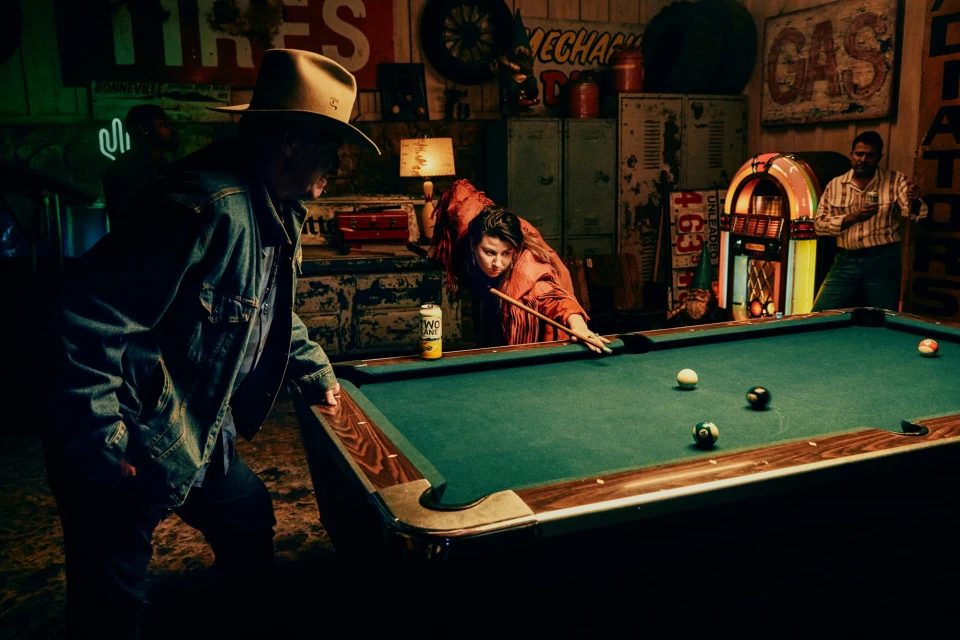 A diverse group gathers around a pool table in a rustic indoor setting, illuminated by a warm, dim light, with a jukebox in the background.