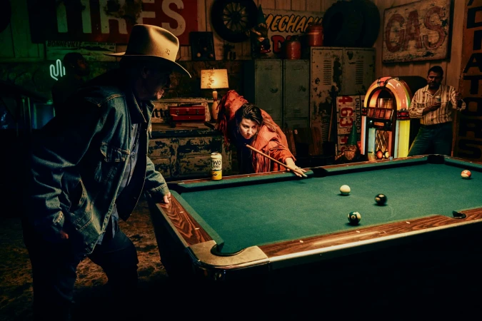 A diverse group gathers around a pool table in a rustic indoor setting, illuminated by a warm, dim light, with a jukebox in the background.