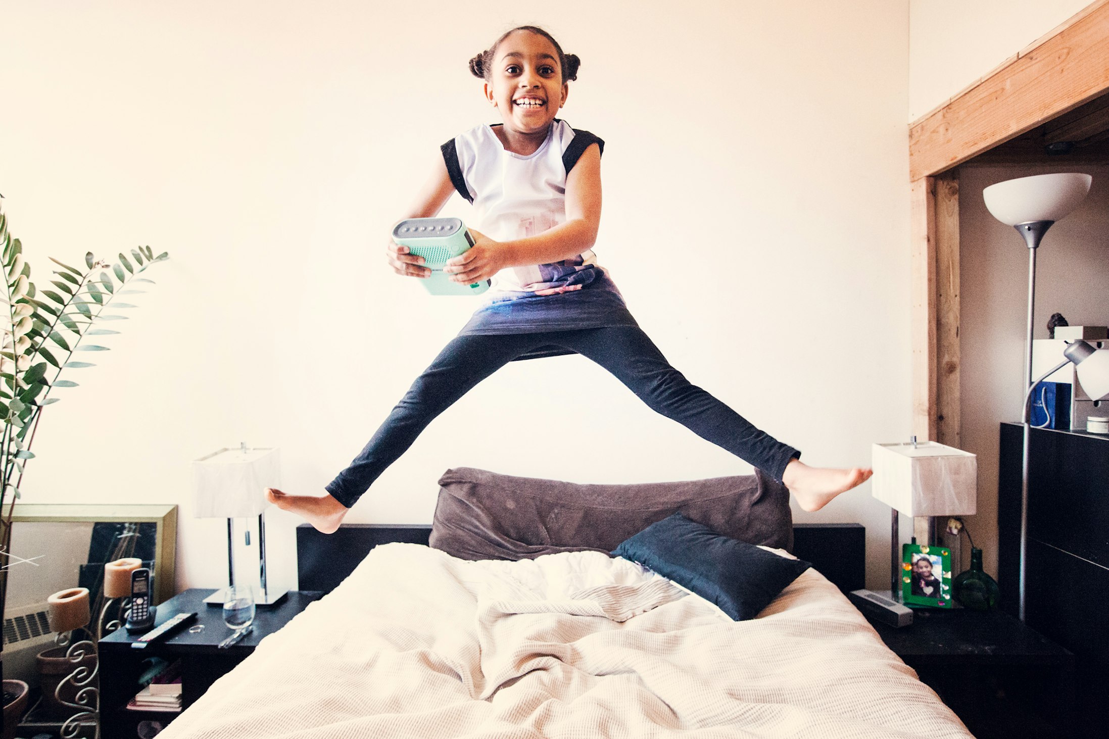 Excited young girl leaps joyfully on a bed in a brightly lit room holding a mint green portable steamer, showing dynamic energy and playfulness.