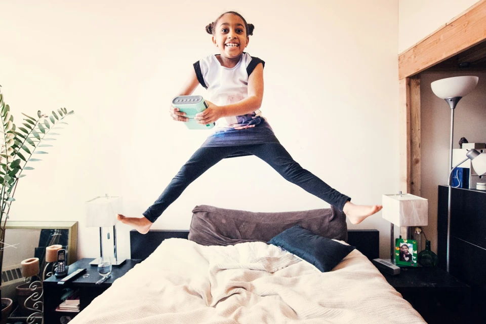Excited young girl leaps joyfully on a bed in a brightly lit room holding a mint green portable steamer, showing dynamic energy and playfulness.