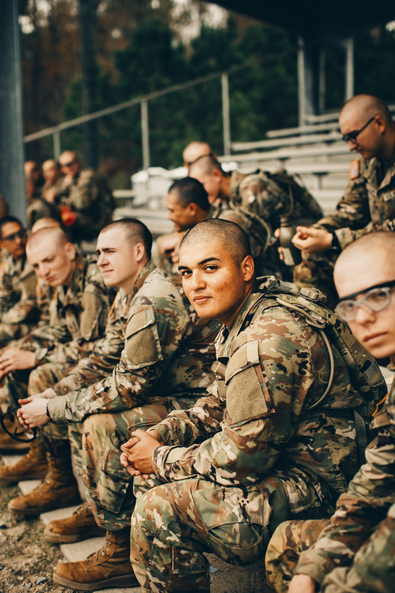 A group of adult male soldiers in camouflage uniforms are seated in bleachers in an outdoor setting with a blurred background.