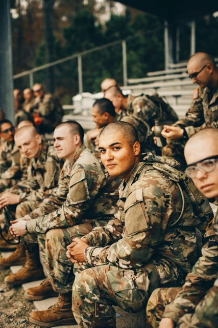 A group of adult male soldiers in camouflage uniforms are seated in bleachers in an outdoor setting with a blurred background.