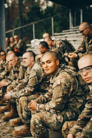 A group of adult male soldiers in camouflage uniforms are seated in bleachers in an outdoor setting with a blurred background.