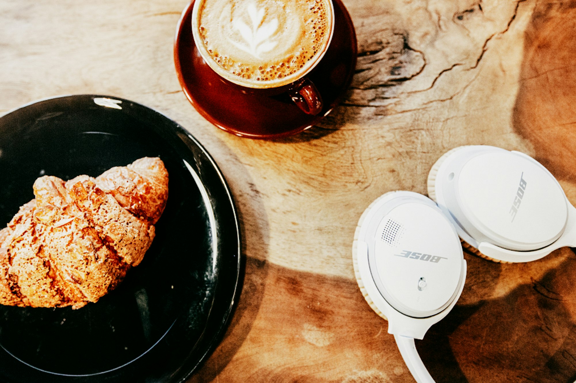 Overhead shot of a cappuccino, croissant, and headphones on a wood table, ideal for a cafe advertisement or tech lifestyle blog.