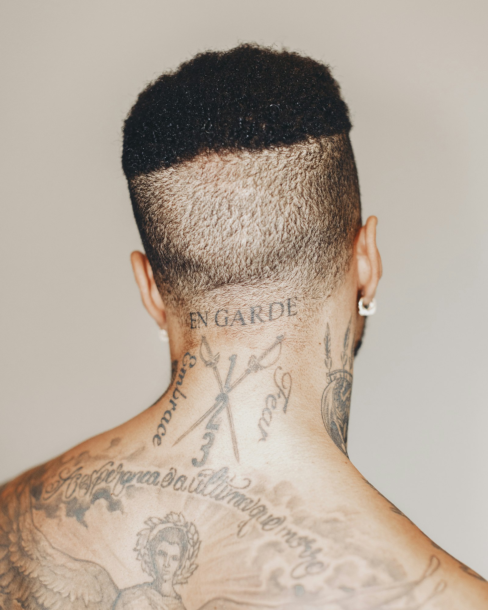 Close-up of a tattooed person's upper back and head showing the back of the head with short, dark, curly hair and multiple tattoos.