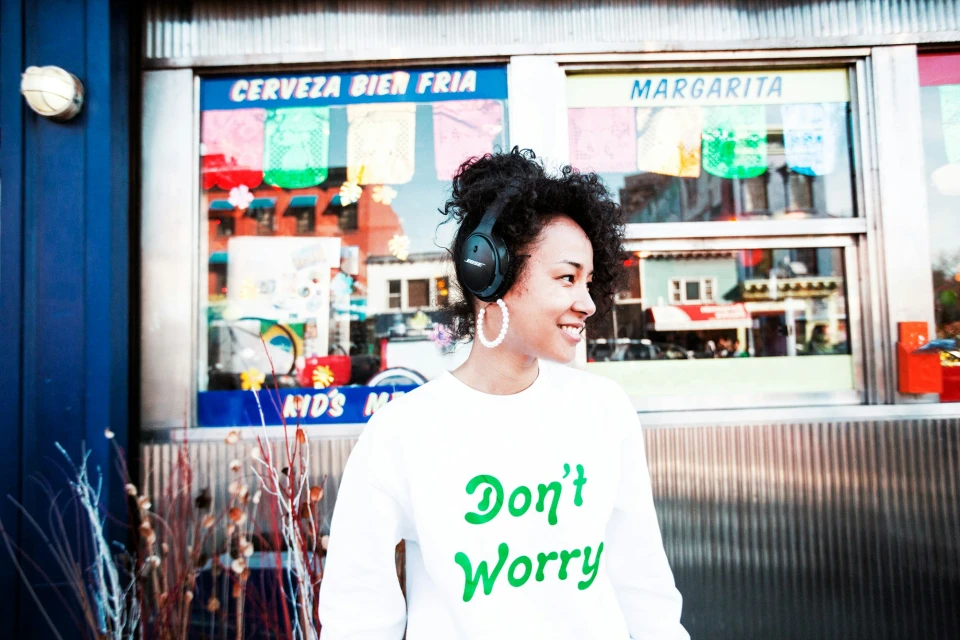 Smiling young woman with afro wearing headphones and a white sweatshirt stands in front of a building with colorful signage.
