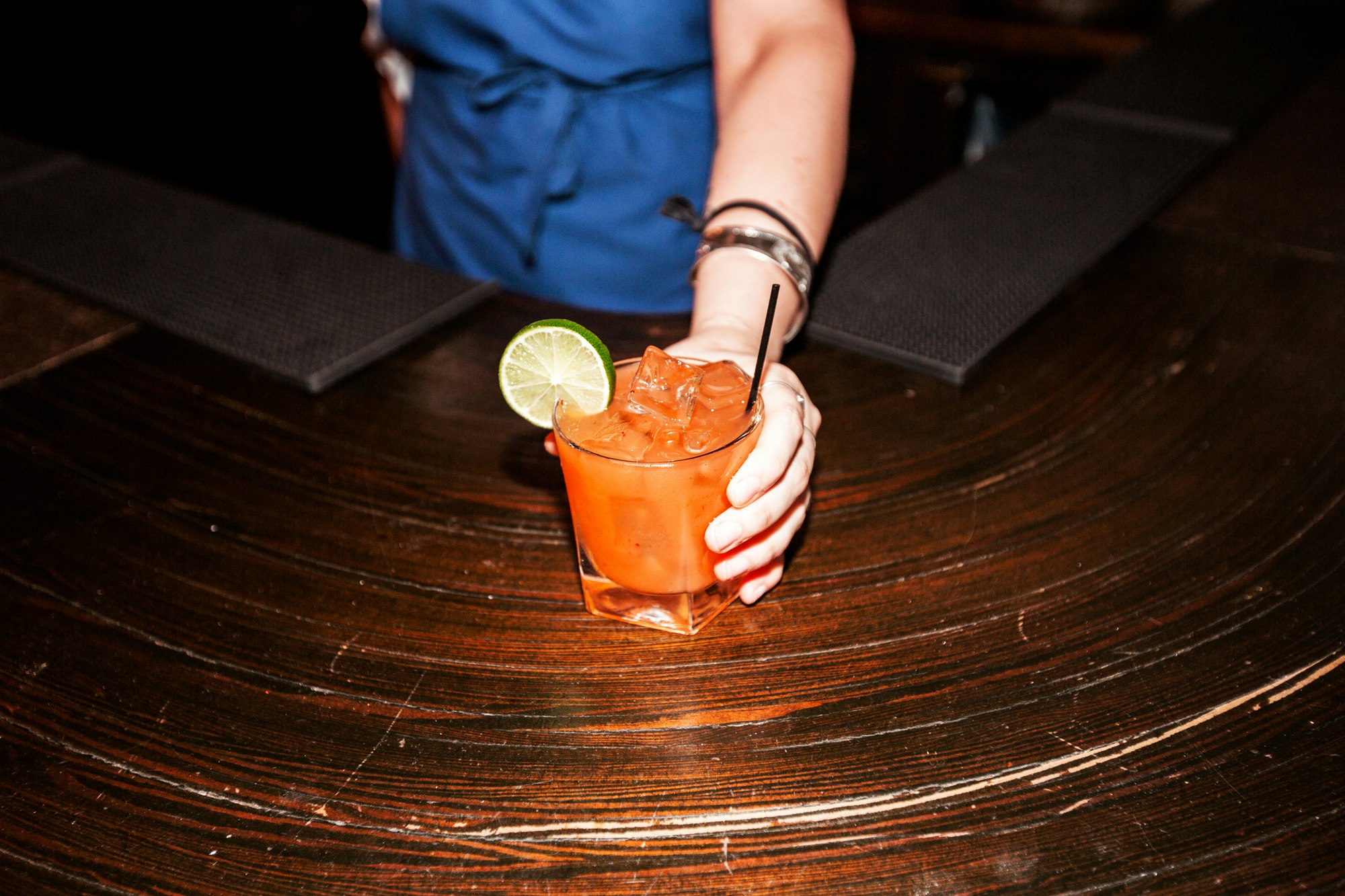 Person's hand holds an orange cocktail with a lime wedge, garnished with a black straw, set on a dark, textured bar top.