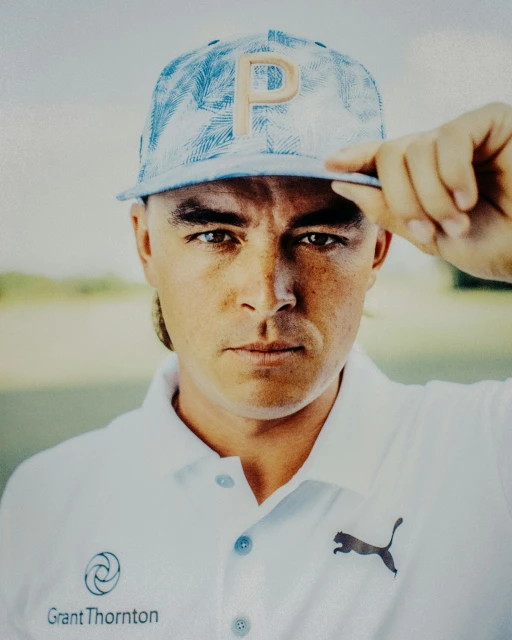Close-up studio shot of an adult male in a white Puma polo shirt and blue patterned cap, posed against a blurry green and white background.