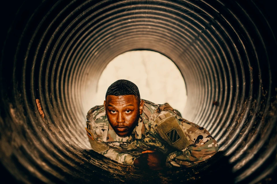Portrait of an adult African-American male in military fatigues, seen inside a metal tunnel, looking directly at the camera.
