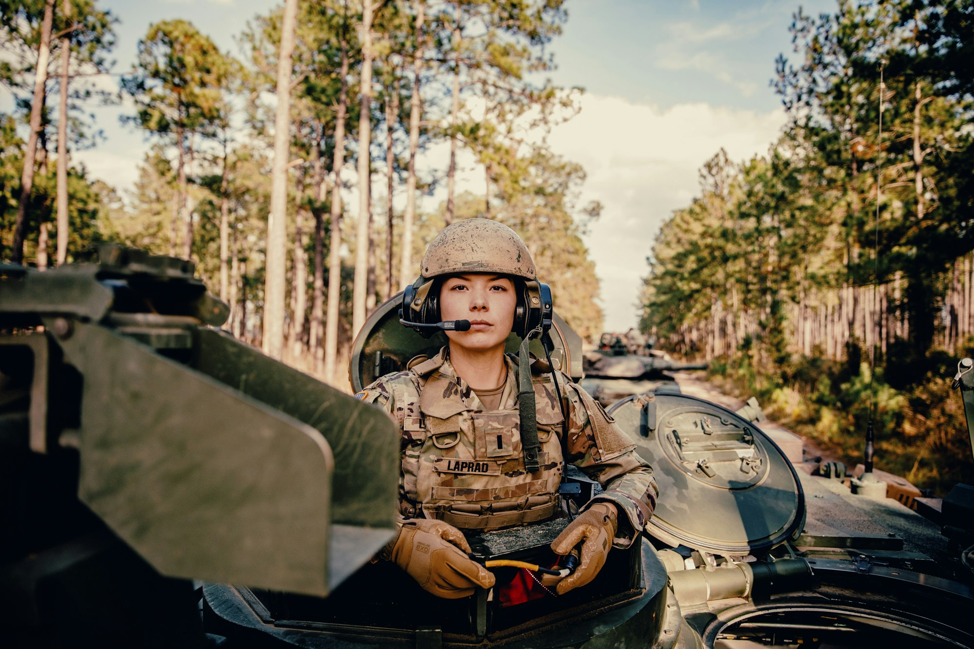 Female soldier in military uniform and helmet sits atop a tank in a forest setting, wearing a headset and gloves.