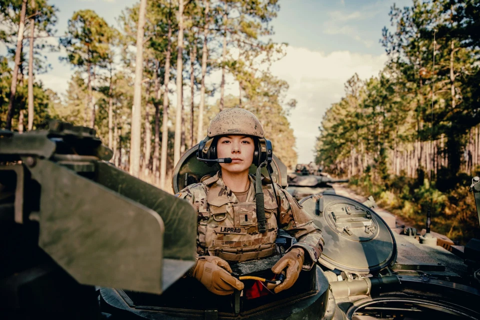 Female soldier in military uniform and helmet sits atop a tank in a forest setting, wearing a headset and gloves.