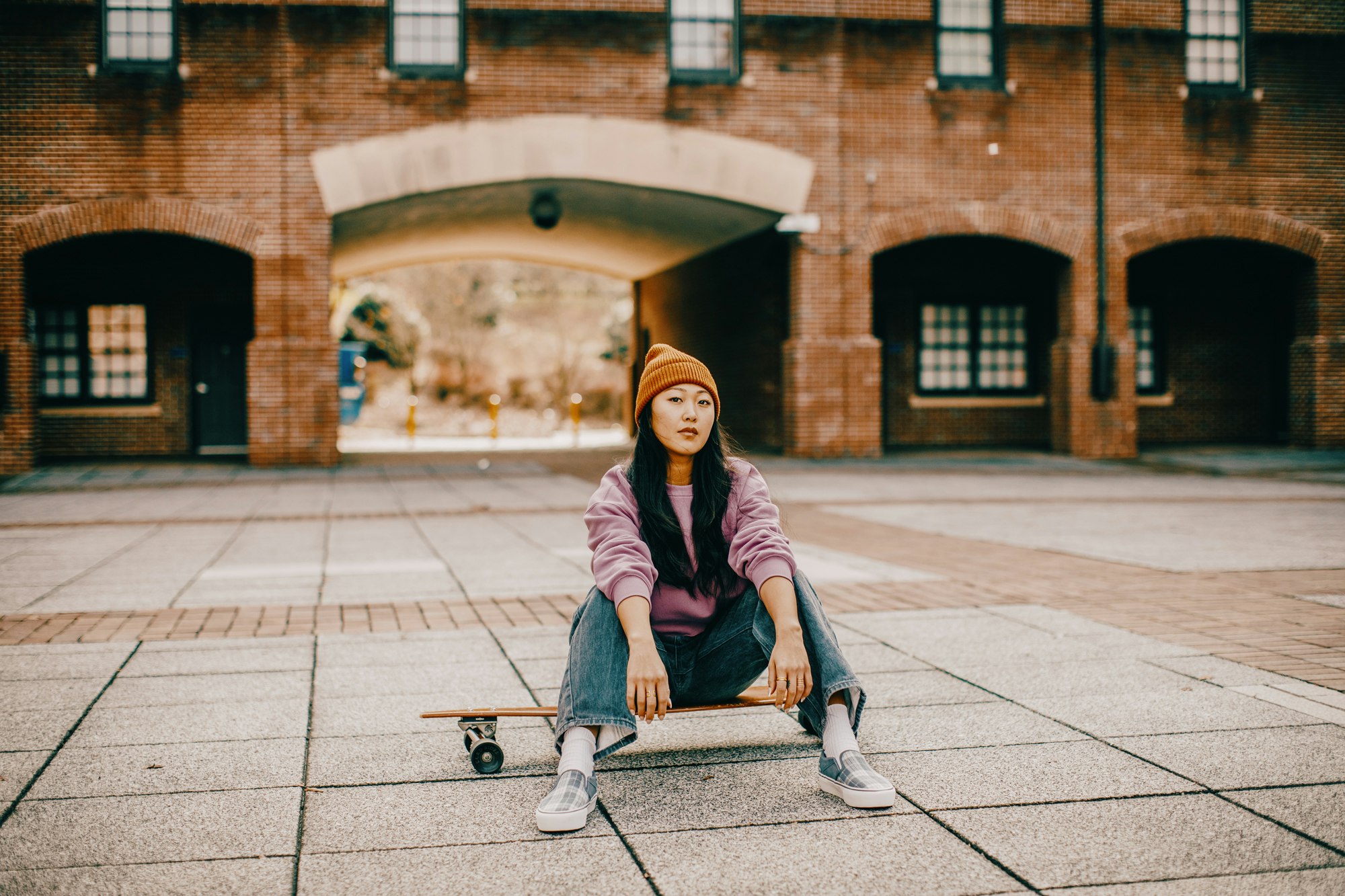 Asian woman with a beanie sits on a skateboard in front of a red brick building, showcasing urban lifestyle and casual fashion.