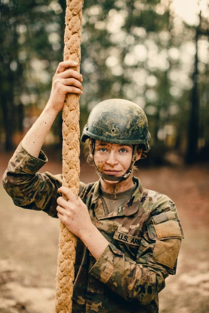Mud-covered female soldier wearing a helmet and camouflage uniform grips a thick rope in a wooded area, looking toward the viewer.