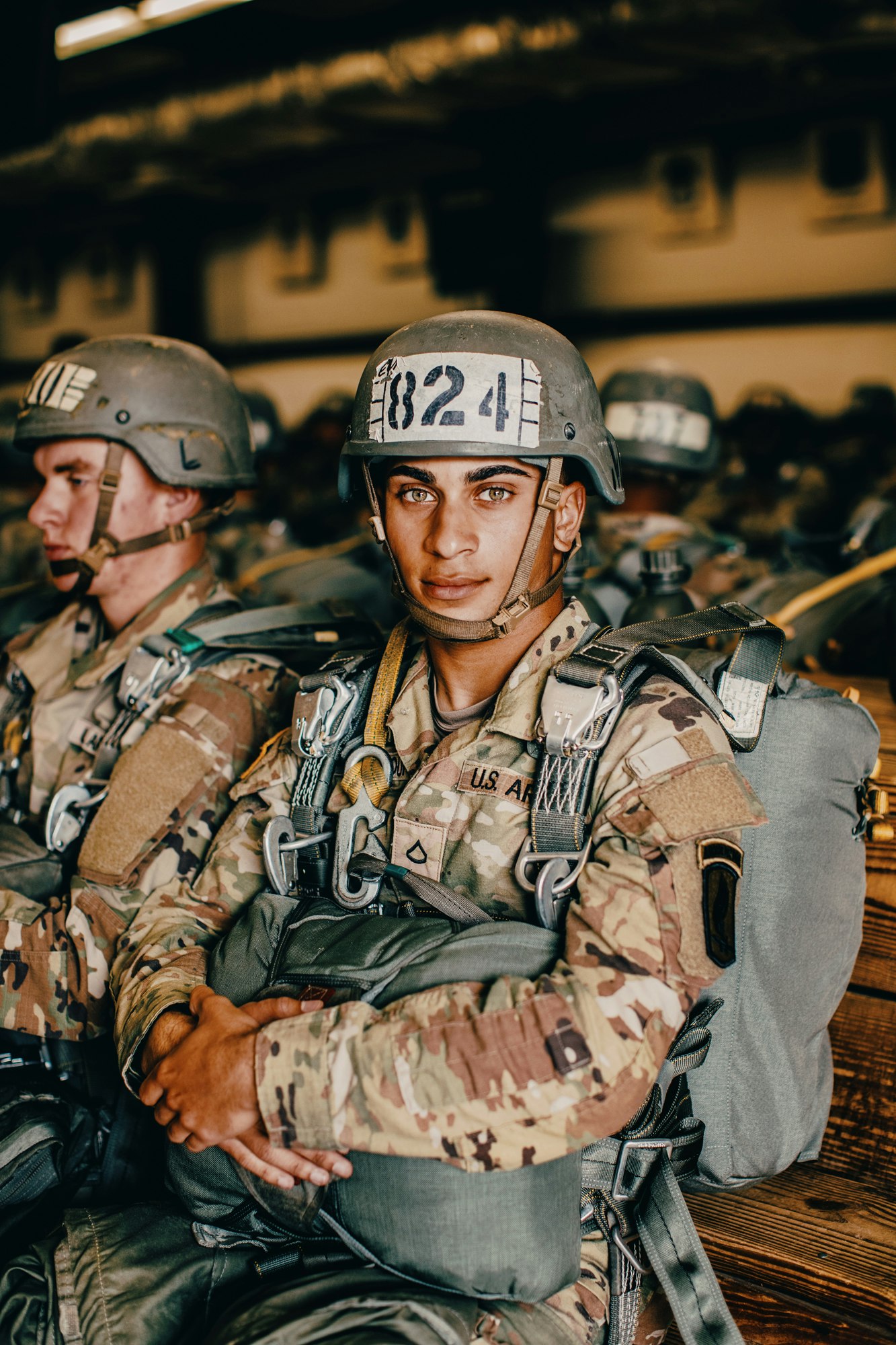 Portrait of a soldier in camouflage uniform wearing a helmet with number 824, seated in an indoor setting with other soldiers.