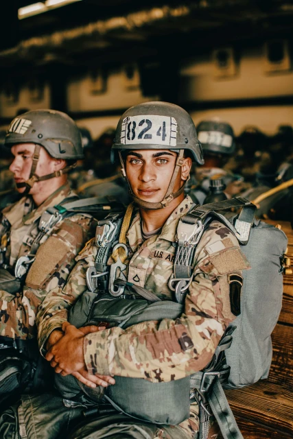 Portrait of a soldier in camouflage uniform wearing a helmet with number 824, seated in an indoor setting with other soldiers.