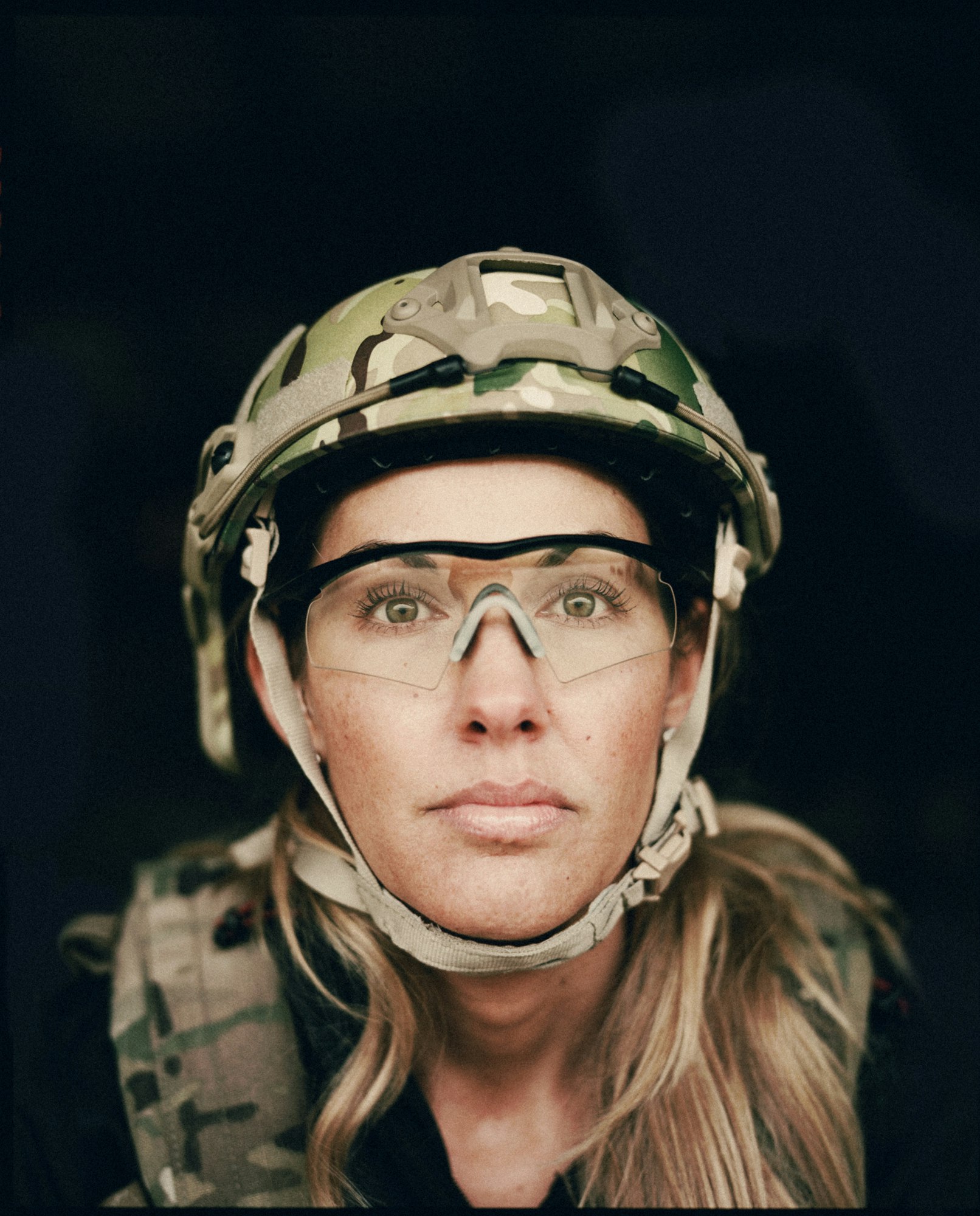 Close-up studio shot of an adult female in a military helmet and vest looking directly at the camera against a black background.