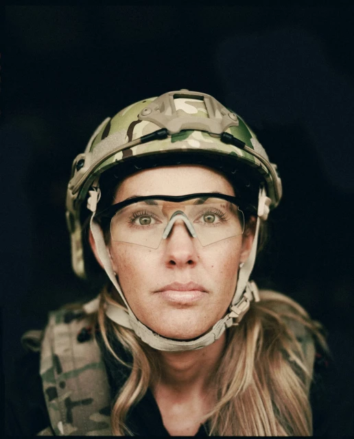 Close-up studio shot of an adult female in a military helmet and vest looking directly at the camera against a black background.