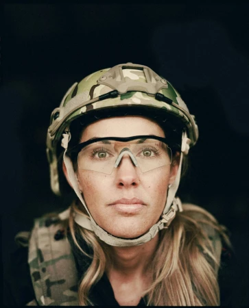 Close-up studio shot of an adult female in a military helmet and vest looking directly at the camera against a black background.