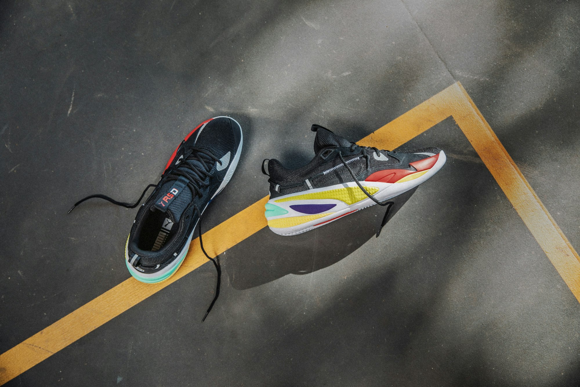 Overhead studio shot of two black Puma RS-D sneakers with colorful soles on a gray surface with painted yellow lines.