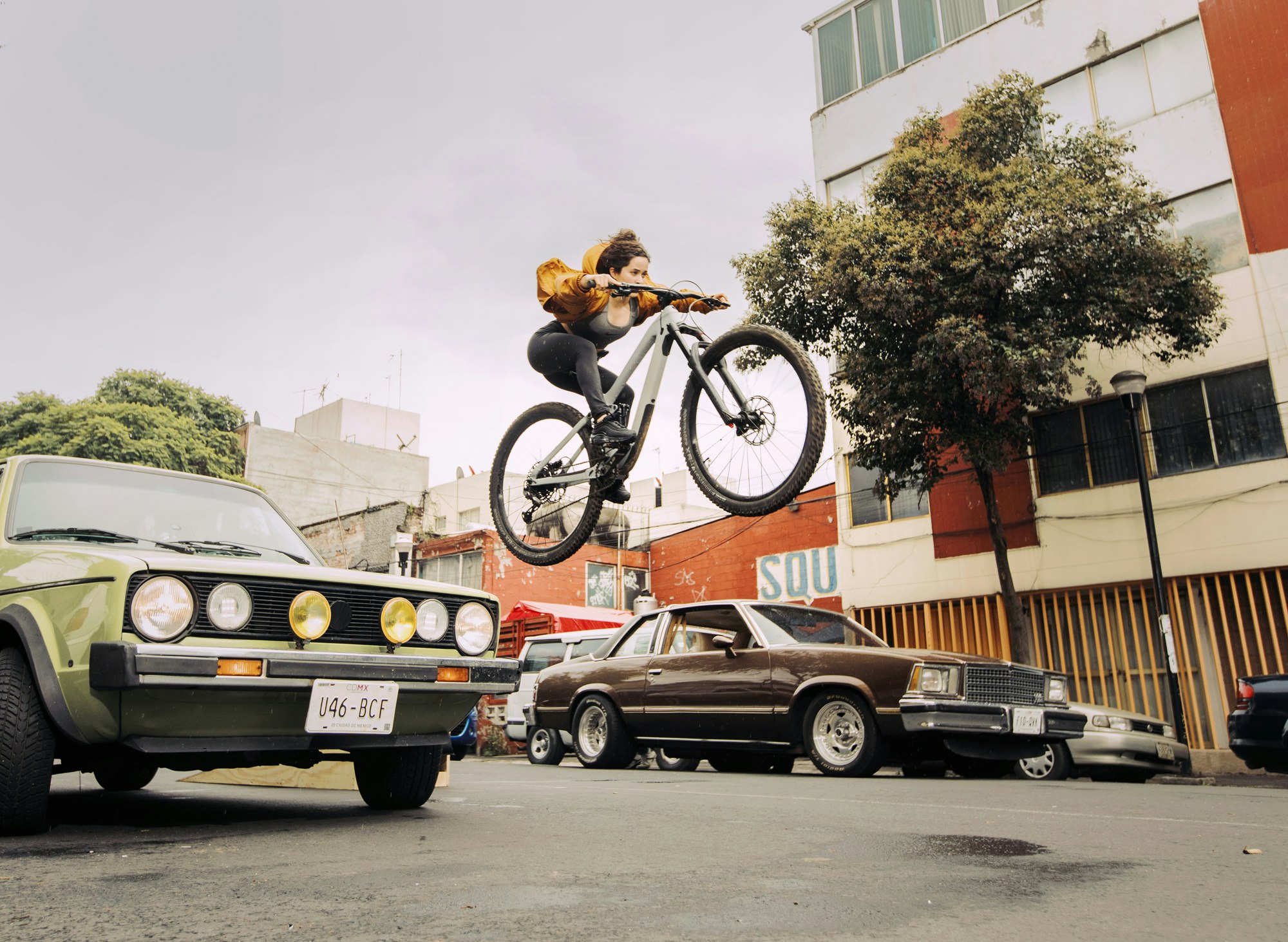 Adult female jumps high on a mountain bike, airborne over a city street with vintage cars and urban buildings, captured in a wide shot.