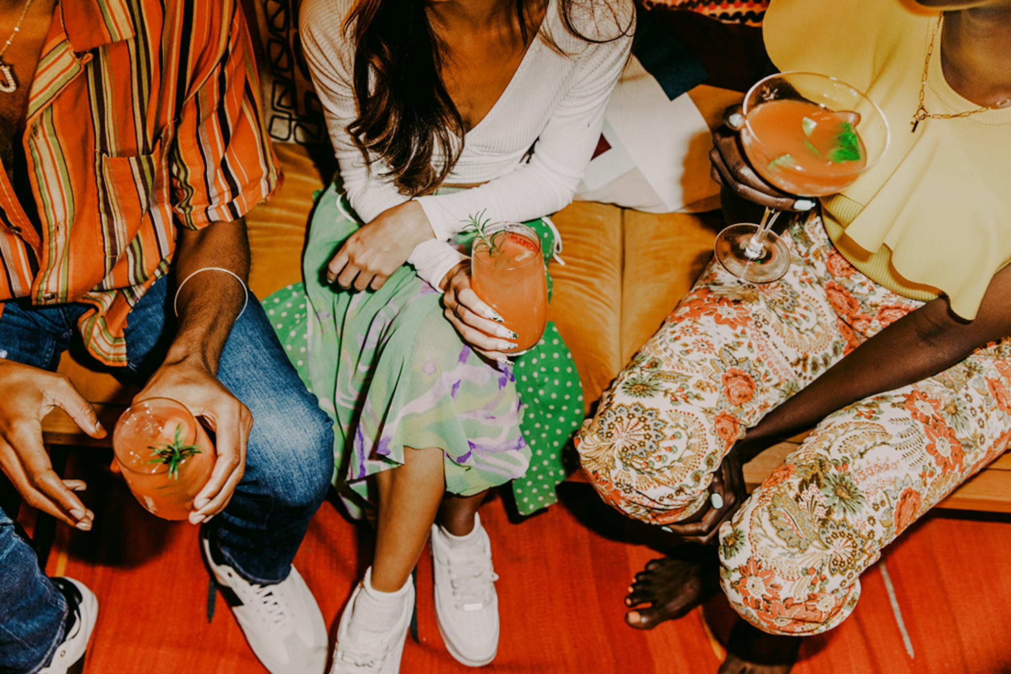 Three friends sit together, holding cocktails in a casual setting. The focus is on their patterned clothing and the vibrant drinks.
