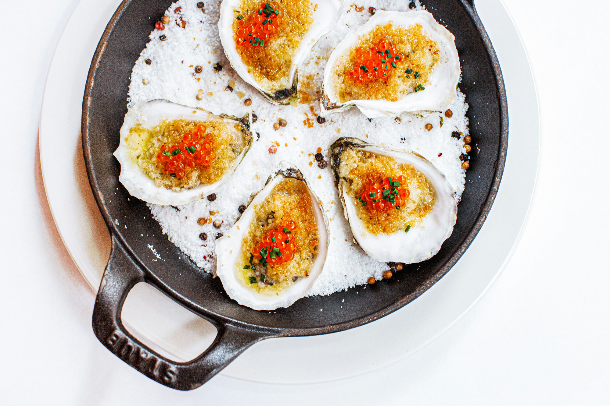 Overhead close-up of five oysters with caviar and herbs on a bed of salt in a cast iron skillet on a white plate.