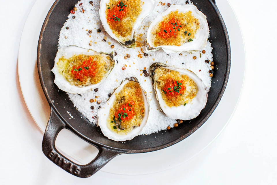 Overhead close-up of five oysters with caviar and herbs on a bed of salt in a cast iron skillet on a white plate.