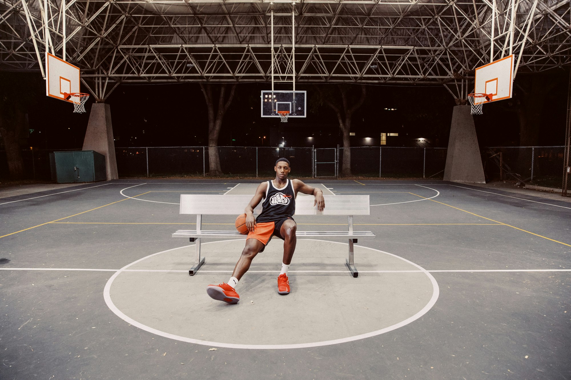 An African American man sits on a bench on an outdoor basketball court at night, holding a basketball and wearing sports clothes.