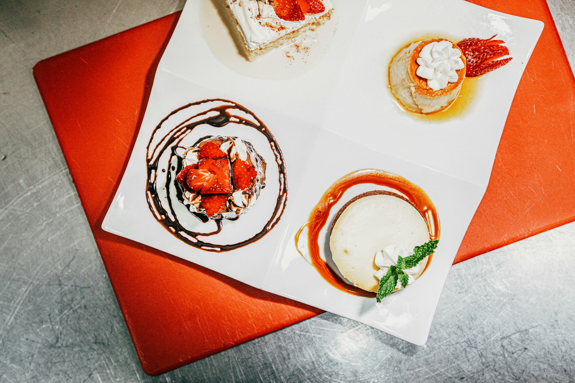 Overhead shot of a white plate holding four desserts: strawberry shortcake, flan, cheesecake, and creme caramel, arranged on a red surface.