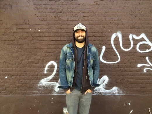 Adult male stands in front of a brown brick wall with graffiti, wearing a denim jacket, hooded sweatshirt, cap, and gray jeans.