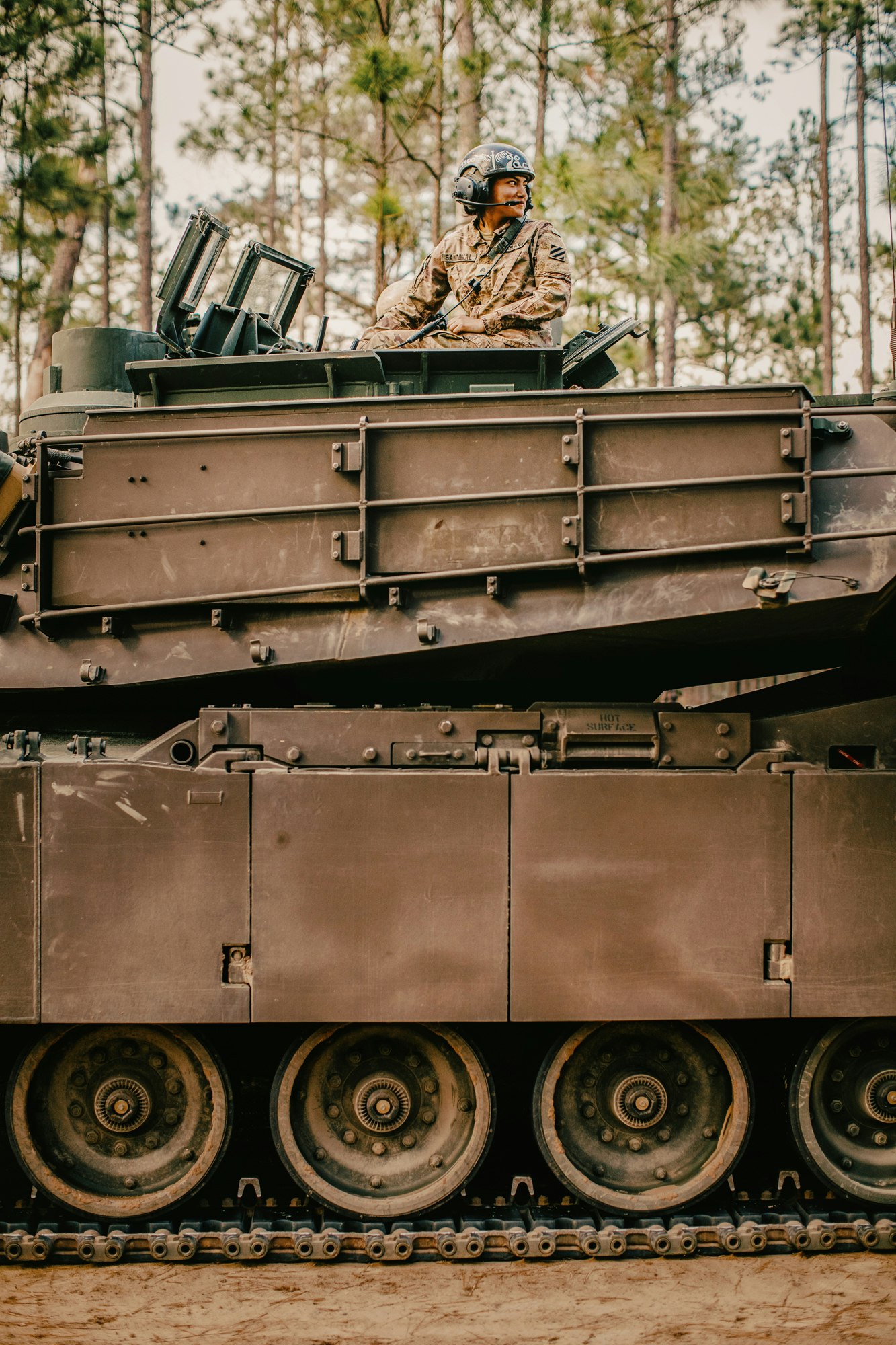 A soldier in camouflage uniform and helmet seated inside the hatch of a military tank outdoors.