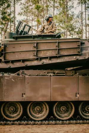 A soldier in camouflage uniform and helmet seated inside the hatch of a military tank outdoors.