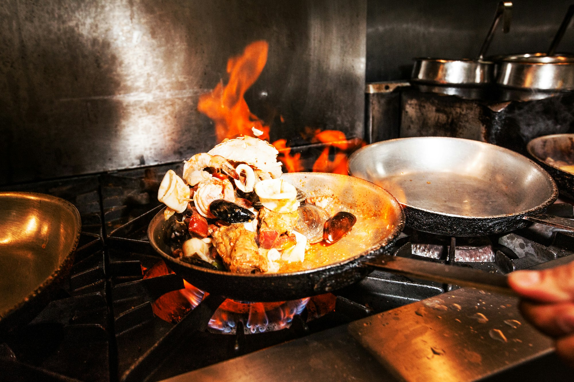 Seafood cioppino cooking in a pan with flames. Mussels, clams, squid, shrimp, and crab simmering on a gas stovetop in a kitchen.
