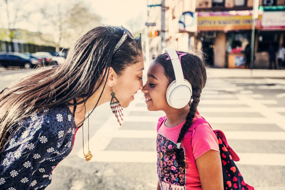 A woman and a girl, possibly mother and daughter, touch foreheads lovingly on a city street. Pedestrians walk on a sunlit crosswalk.