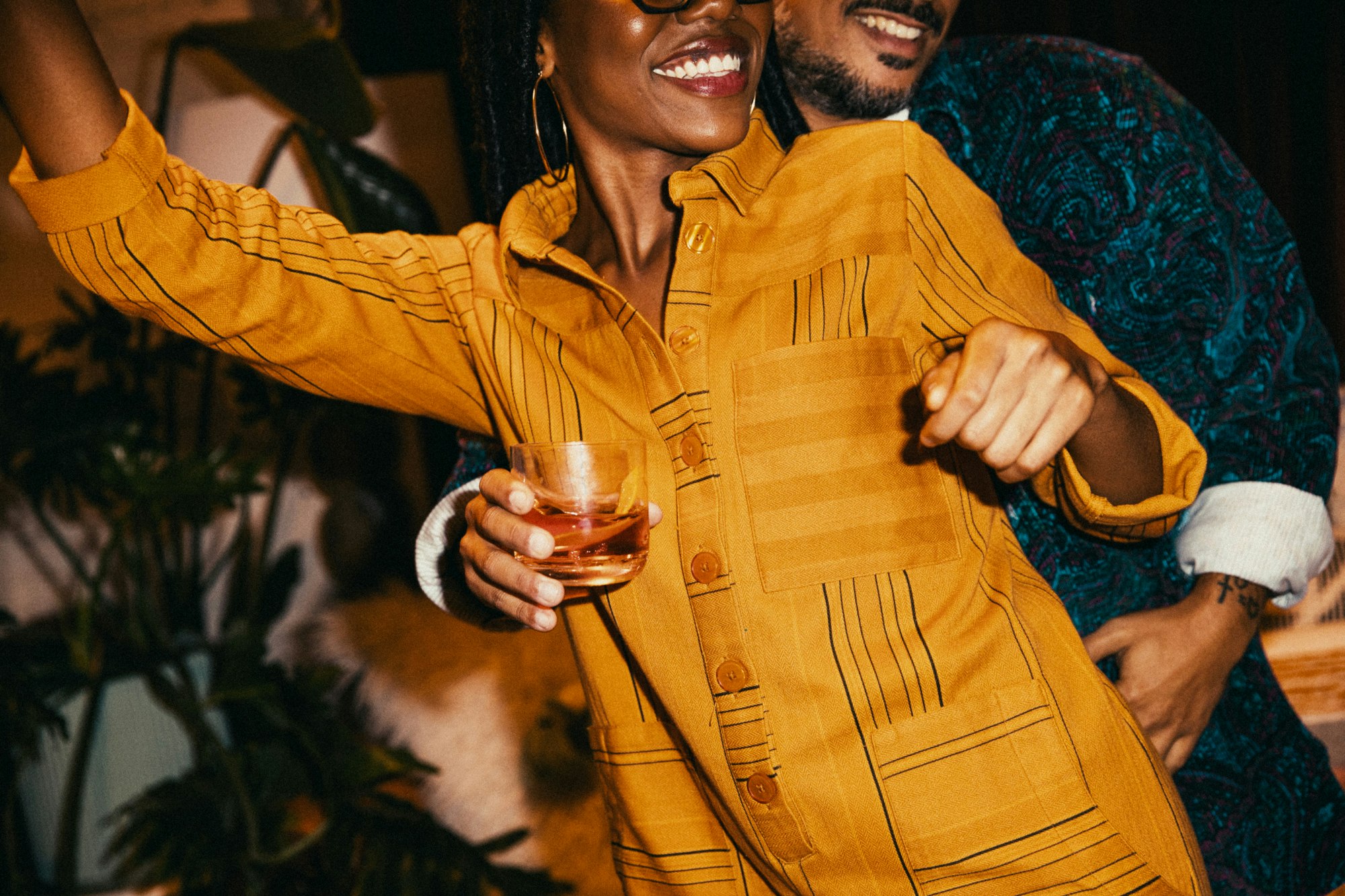 A smiling woman in a yellow shirt dress holds a drink at an indoor party, embracing a man in a paisley jacket, amid indoor plants.