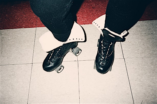 Close-up of roller skates with black leggings and white cuffs on a tiled floor with a red and gray border in the background.