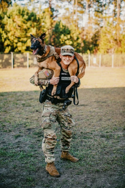 Man in military uniform carries a dog on his shoulders outdoors. The dog is wearing a vest, and the man is wearing a police vest.