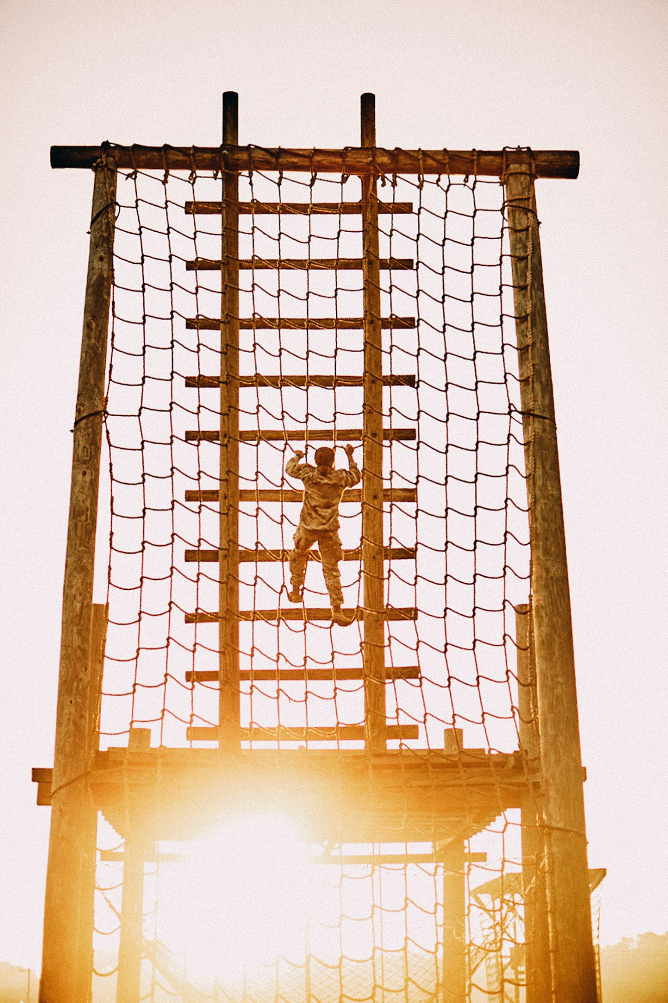 Adult climbs up a wooden and rope ladder structure, silhouetted against the bright, warm light of the sun, in a medium outdoor shot.