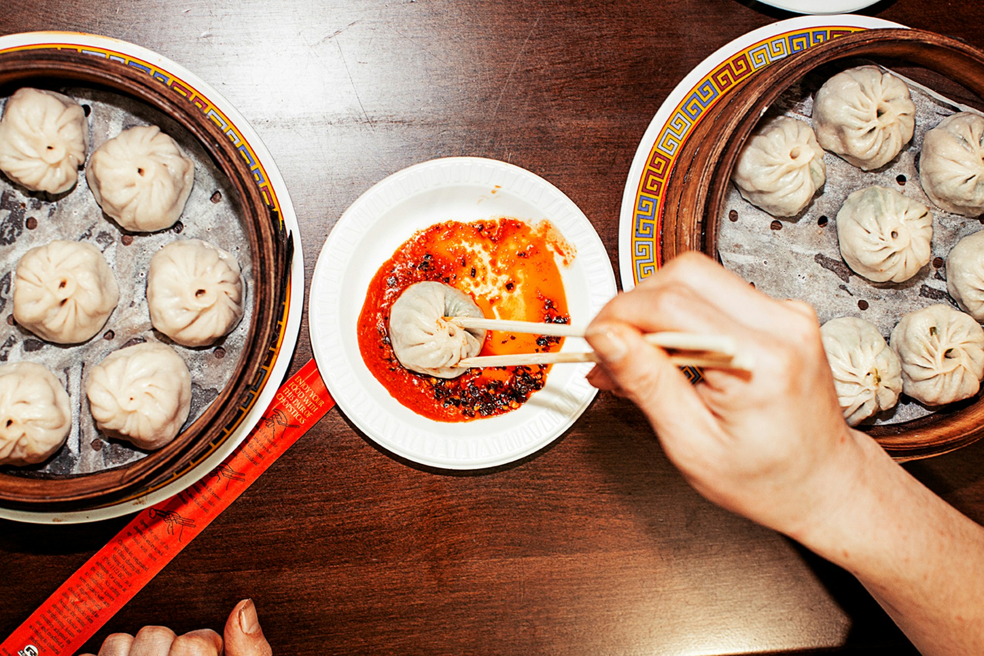 Overhead shot of two bamboo steamers filled with dumplings, and a hand using chopsticks to pick up a dumpling with red sauce.