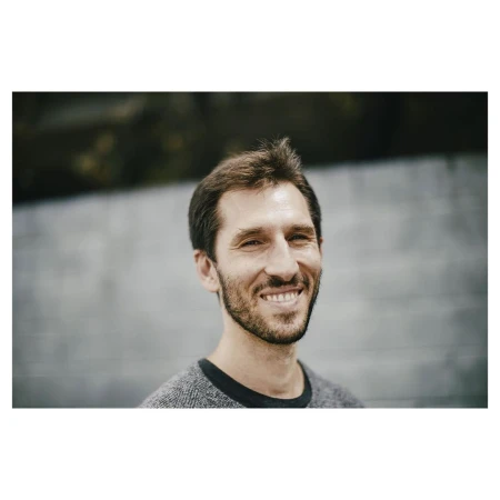 Portrait of a smiling man with short brown hair and beard wearing a grey sweater against a blurred wall
