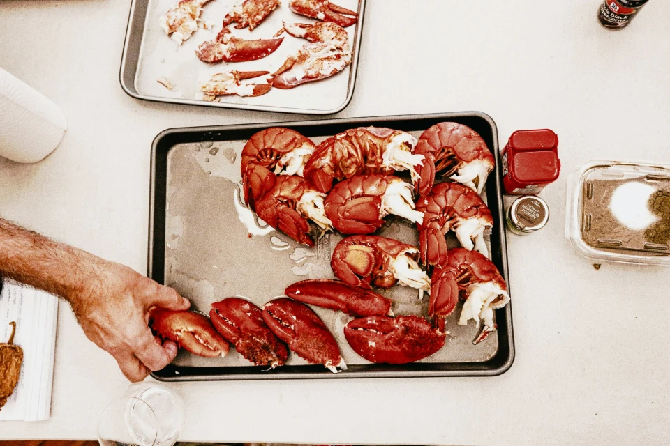 Overhead shot of cooked lobster tails and claws on a metal tray, ready to be served, alongside spices and a beverage.