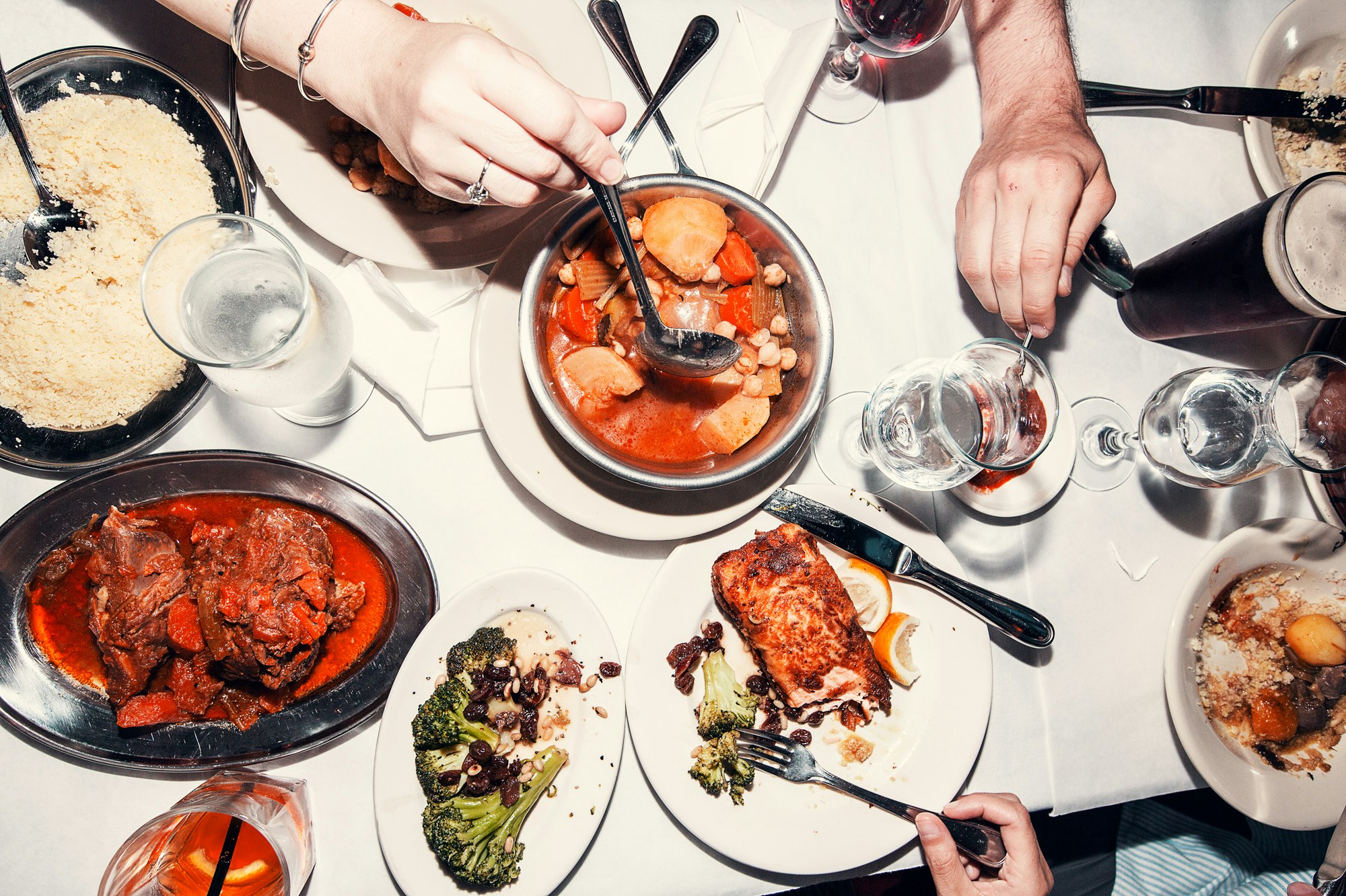 Overhead shot of people dining at a table laden with various dishes, including stew, salmon, vegetables, and couscous, along with drinks.