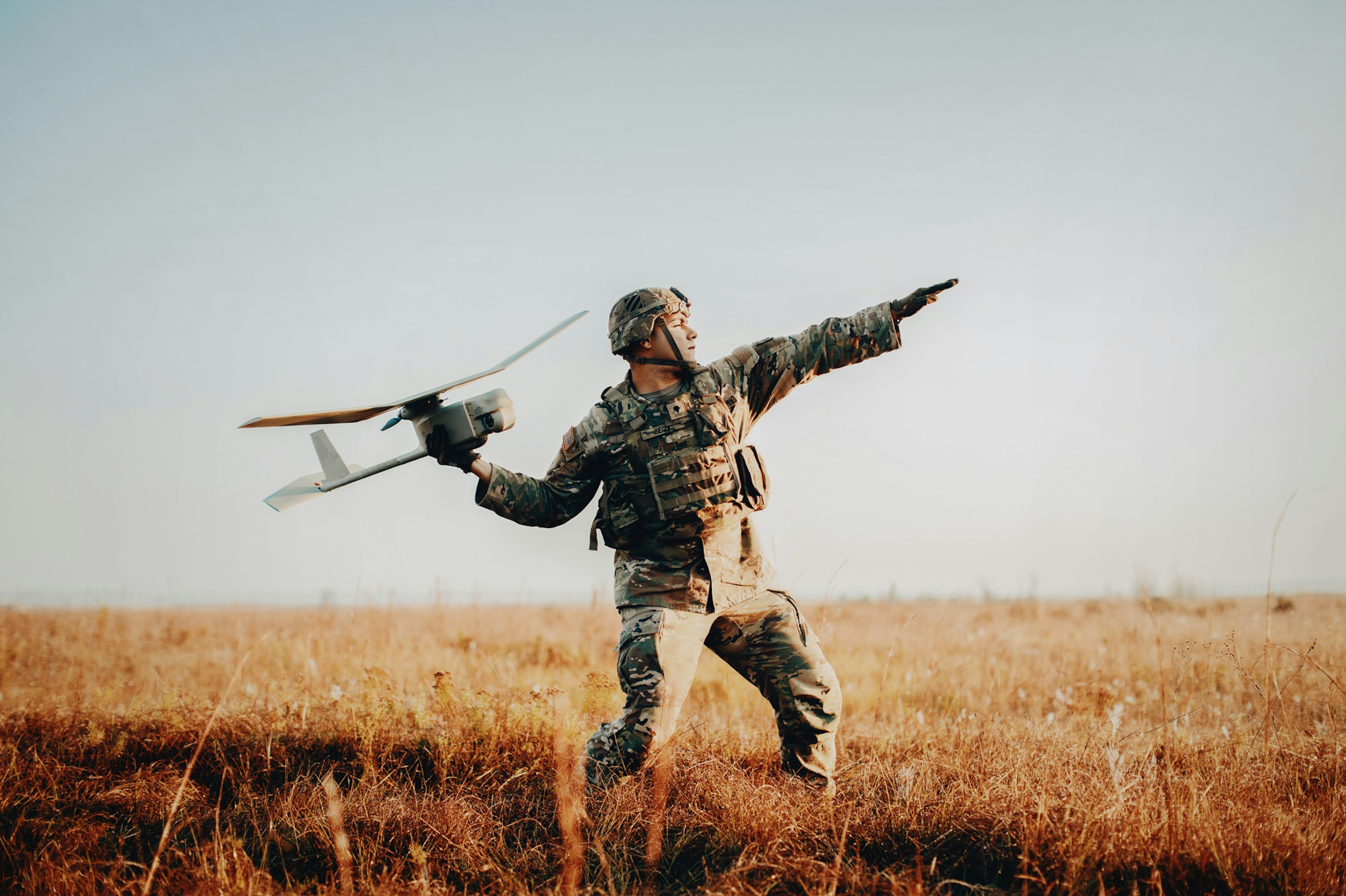 Adult male soldier in camouflage uniform prepares to launch a small drone in a rural, grassy field under a clear sky.