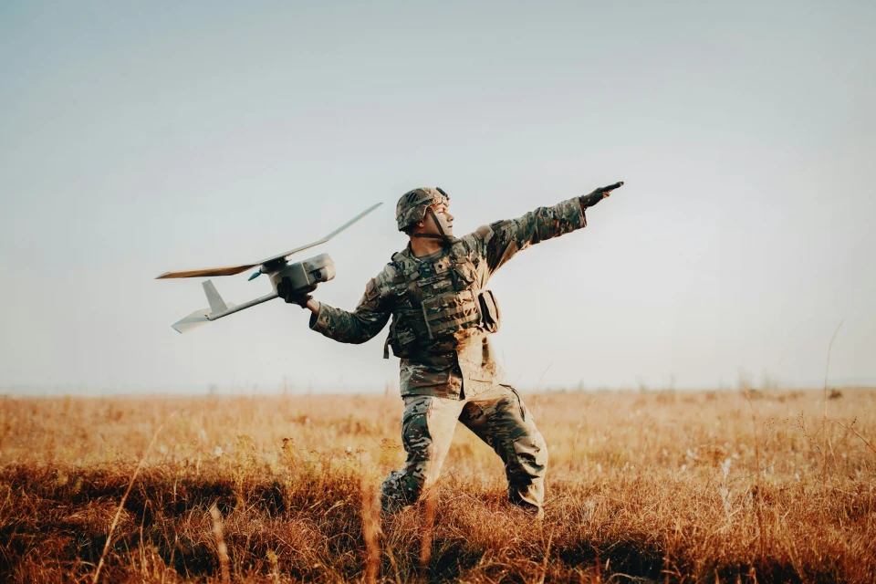Adult male soldier in camouflage uniform prepares to launch a small drone in a rural, grassy field under a clear sky.