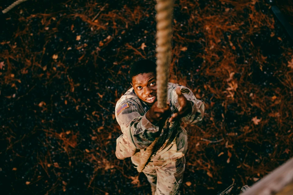 Low angle view of determined African-American adult male in camouflage uniform climbing rope in an outdoor wooded environment.