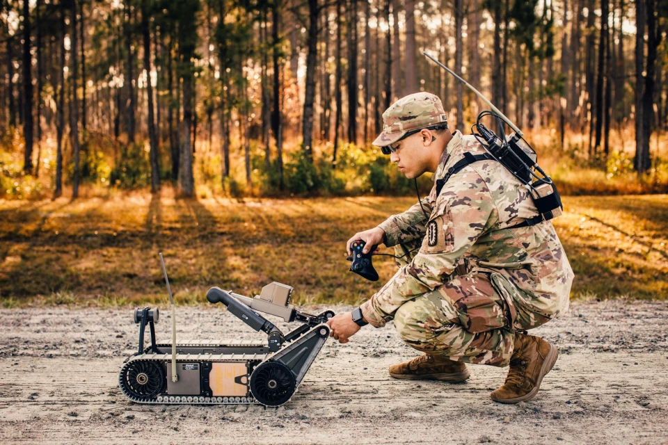 Man in military camouflage operates a tracked robot outdoors in a forest, wearing glasses and gear with trees in the background.