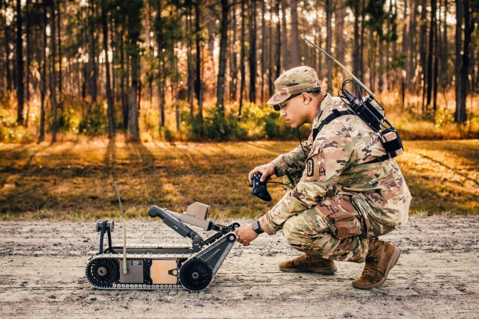 Man in military camouflage operates a tracked robot outdoors in a forest, wearing glasses and gear with trees in the background.