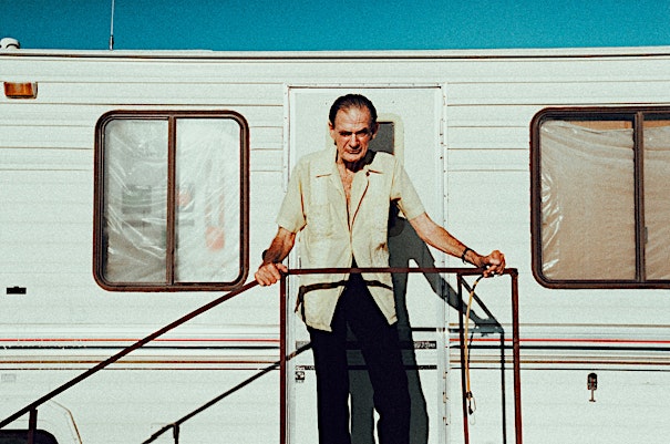 Elderly man stands on the landing of a white recreational vehicle against a clear blue sky. He is wearing a tan shirt and black pants.